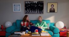 Two women sit on a bright blue couch in a sunny living room.